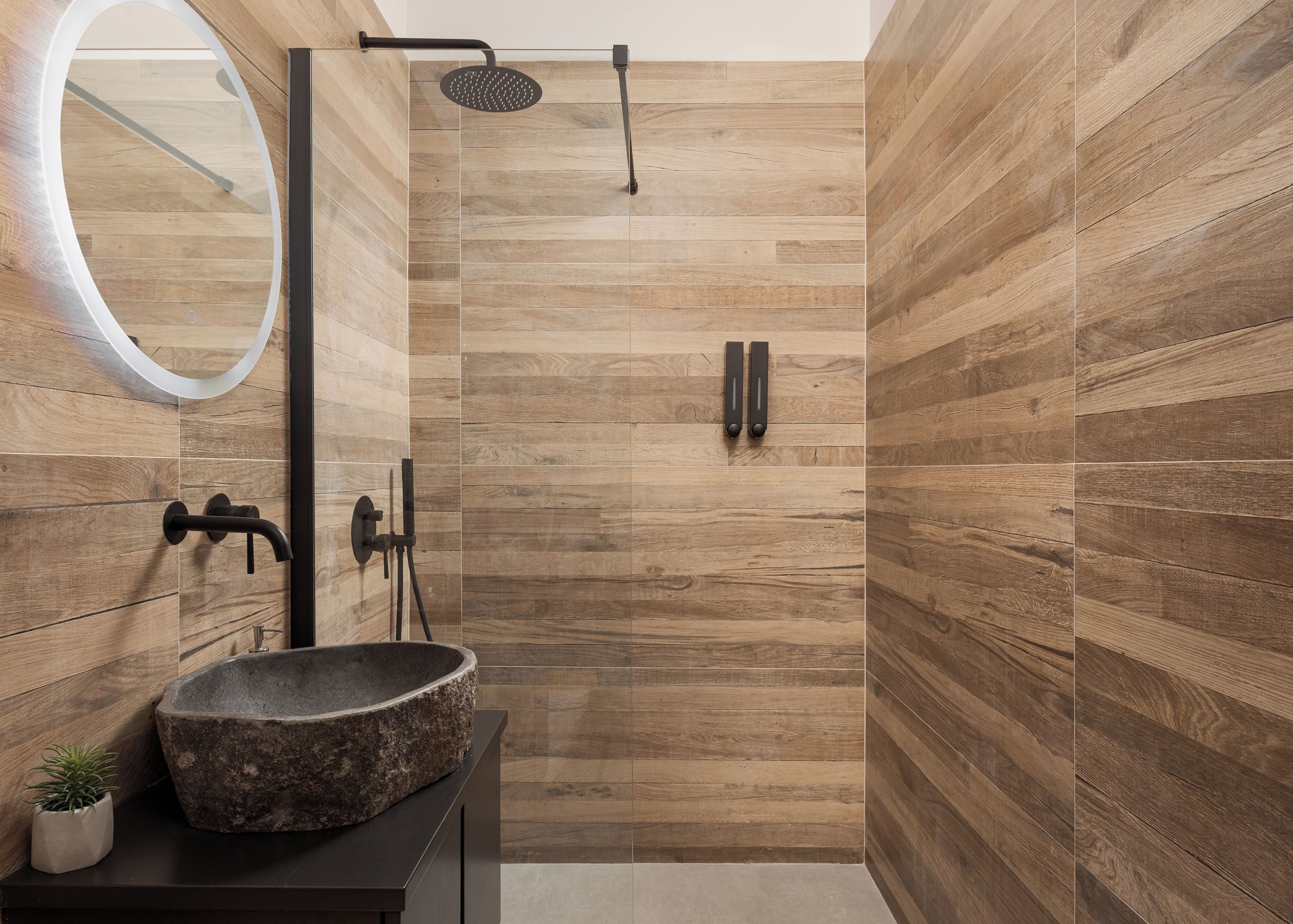 Un baño moderno con pared de madera, un espejo redondo, lavamanos de piedra y ducha con cabezal de ducha manual.