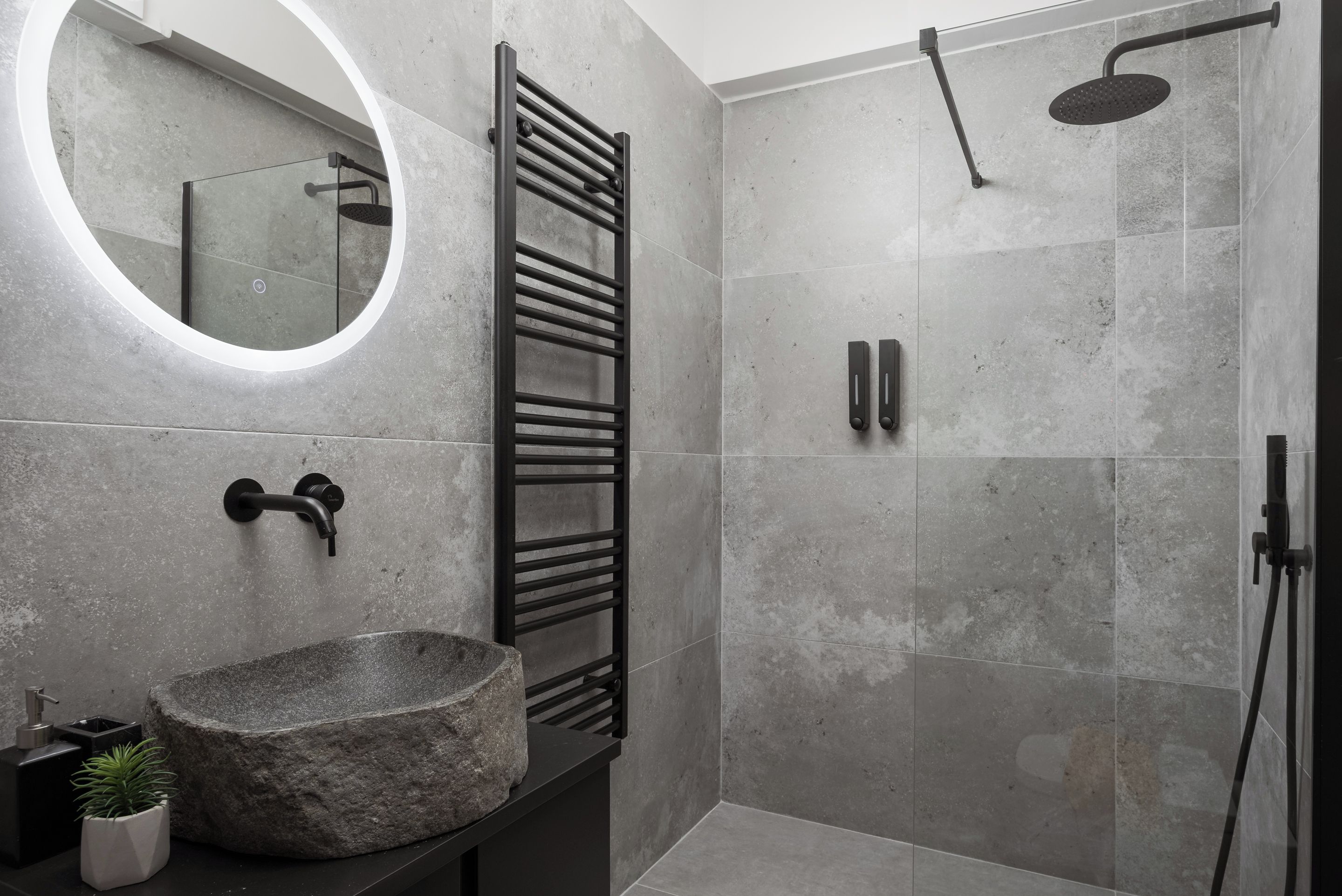 A modern bathroom with a stone sink, a towel rack, a shower with a handheld showerhead, and a round mirror.