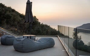 A modern outdoor seating area with a glass balustrade overlooking a body of water and hills. - The One Limeni (Areopoli)