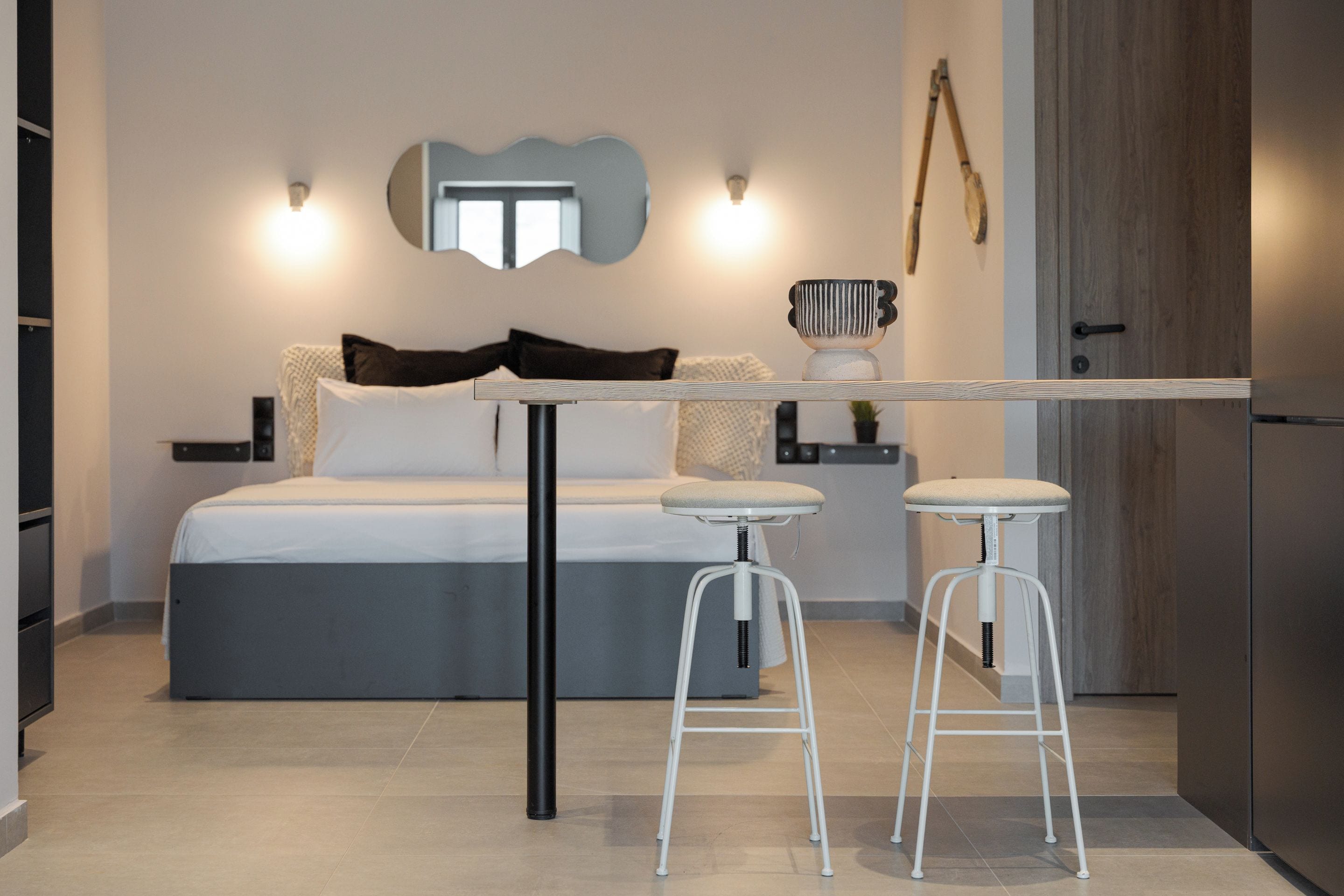 A modern bedroom with a bed, two white bar stools, a small table, and a mirror on the wall.