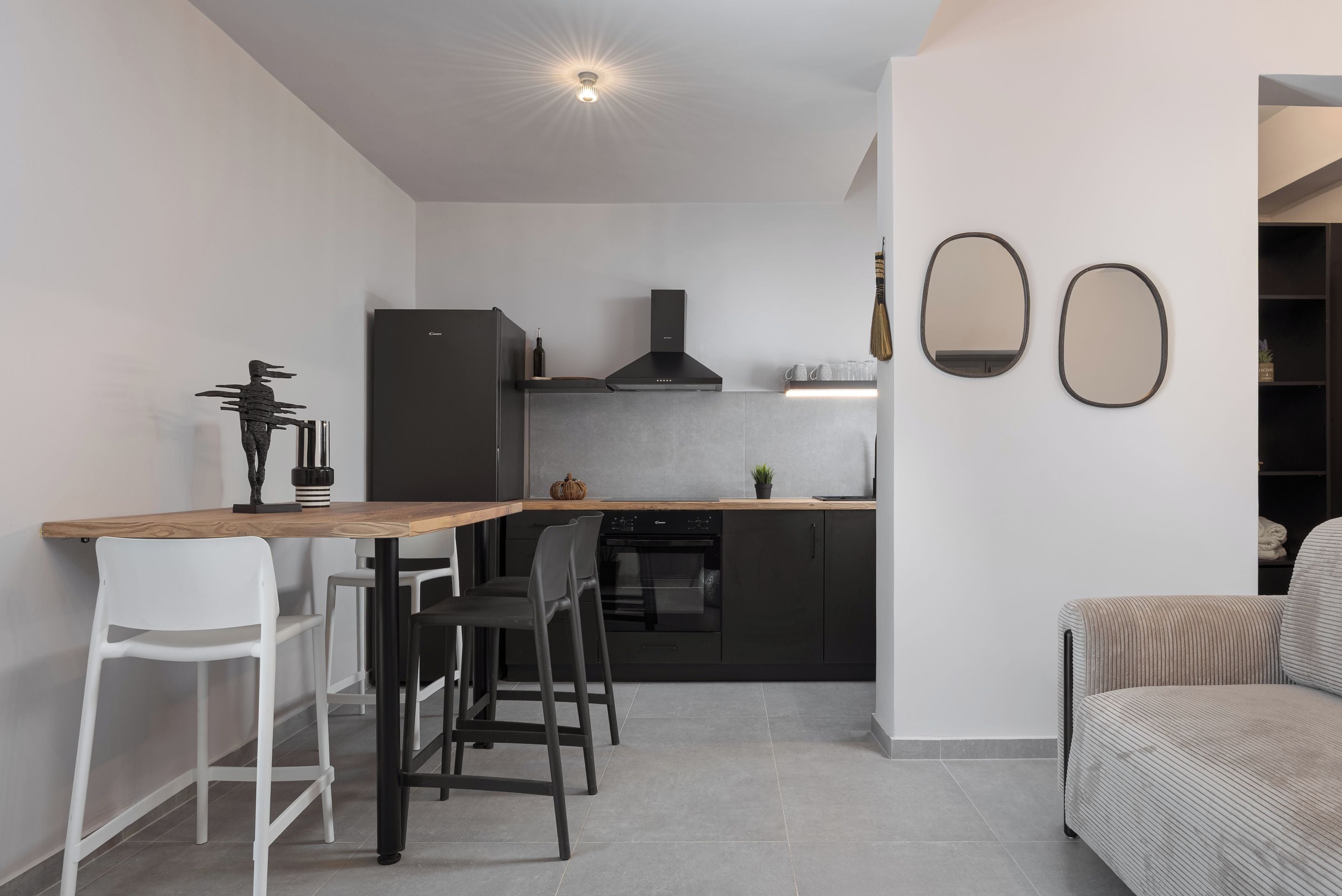 Una cocina moderna con encimera de madera, armarios negros y horno empotrado. En la zona de estar contigua, hay dos taburetes blancos y un sofá gris.