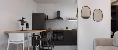 Une cuisine moderne avec un plan de travail en bois, des armoires noires et un four encastré. On y trouve deux tabourets de bar blancs et un canapé gris dans le salon attenant.