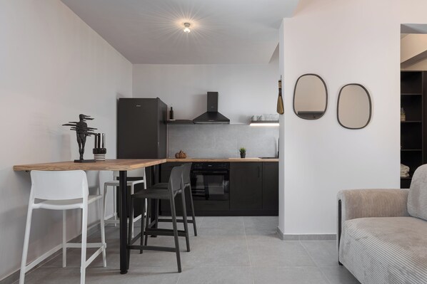 Una cocina moderna con encimera de madera, armarios negros y horno empotrado. En la zona de estar contigua, hay dos taburetes blancos y un sofá gris.