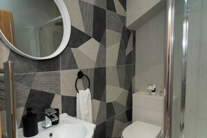 Un bagno moderno con un pannello decorativo a mosaico geometrico, uno specchio rotondo, un lavandino bianco, un water e uno spazio doccia in vetro.
