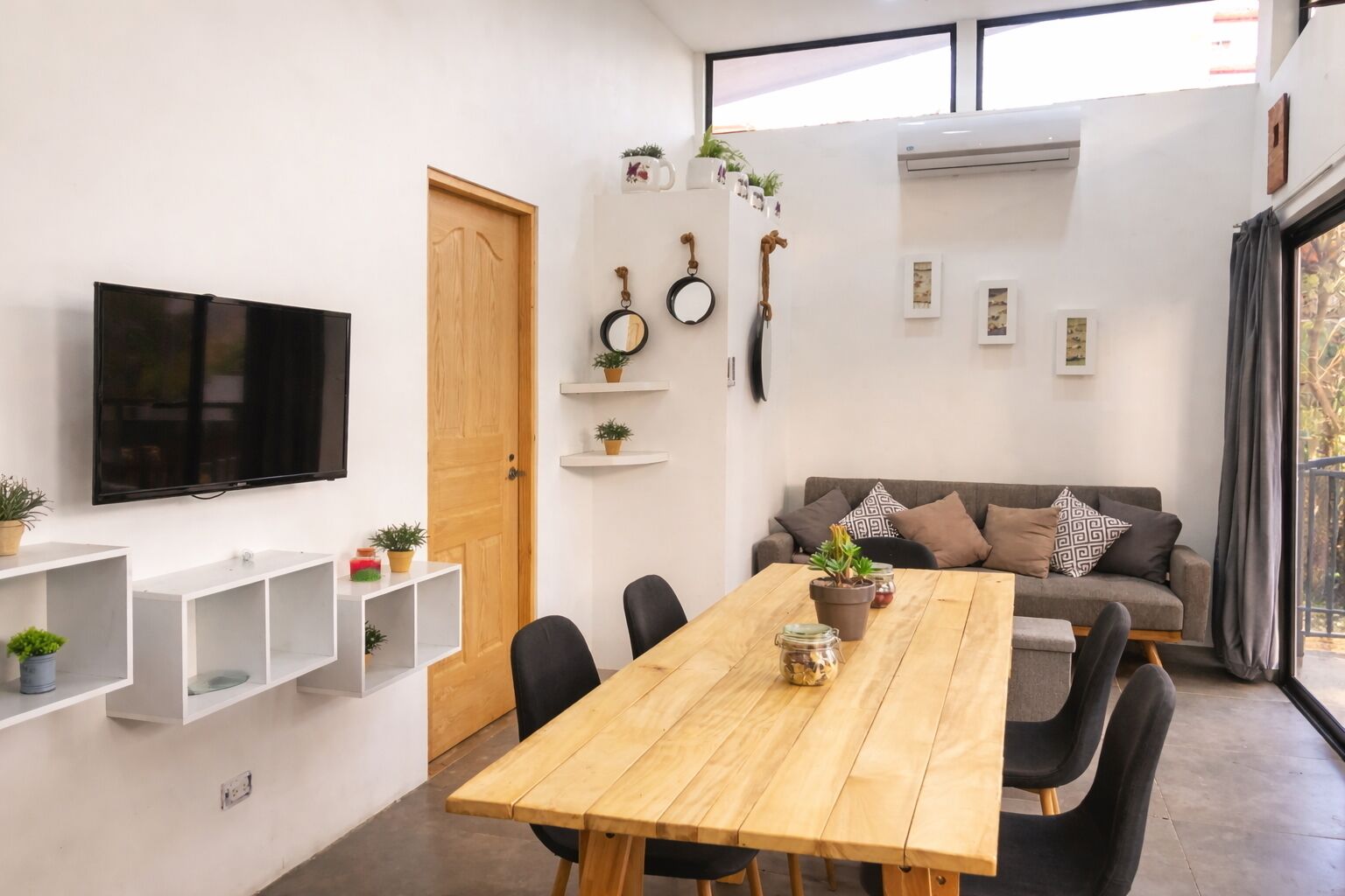 A modern living room with a wooden dining table, black chairs, a grey sofa, and a mounted TV.