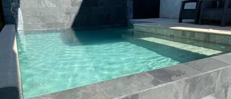 Ein rechteckiger Pool mit klarem Wasser, umgeben von Steinplatten und einer Sitzgelegenheit mit Stühlen.