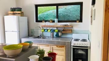 A compact kitchen with a refrigerator, stove, sink, and window with a view of greenery.