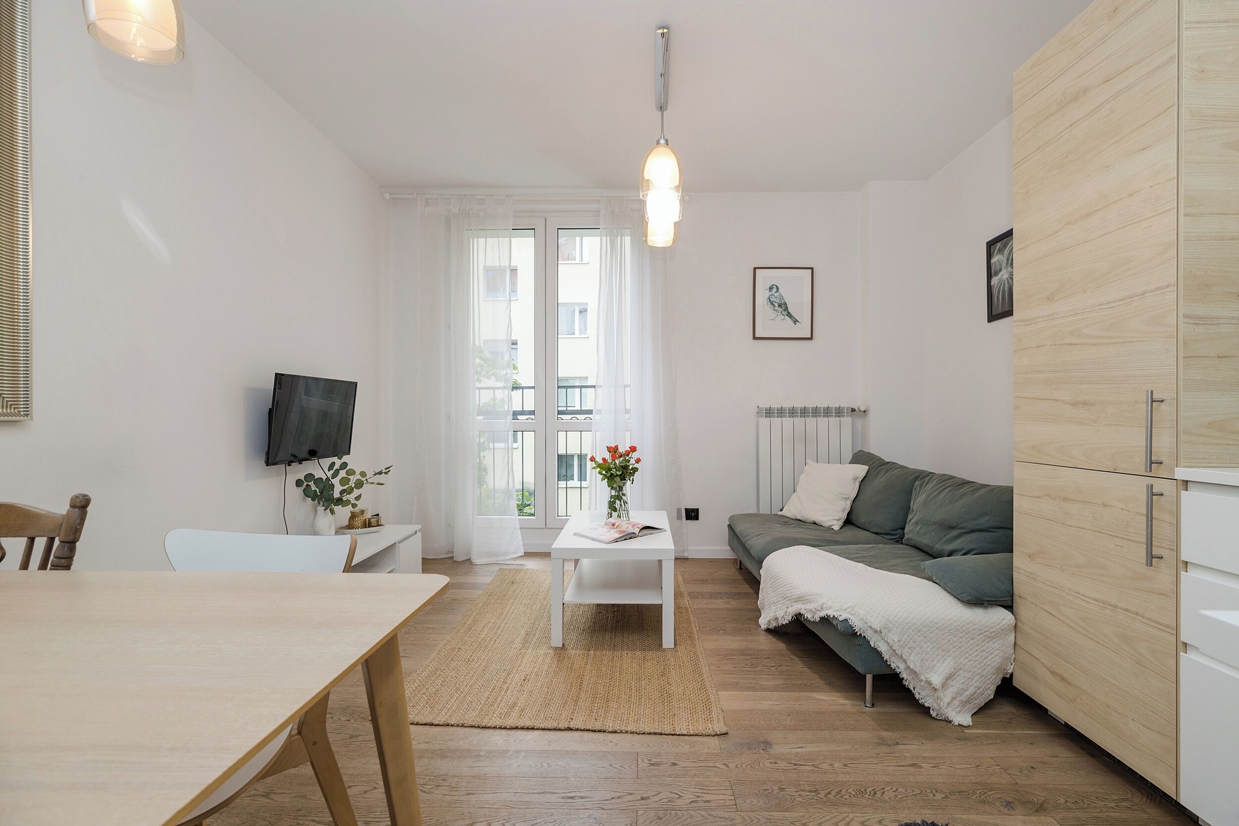 Een moderne woonkamer met een houten vloer, een witte bank, een eettafel en een aan de muur bevestigde televisie.
