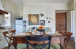 Un coin repas avec une table en bois, des chaises, un tableau au mur et une cuisine en arrière-plan.