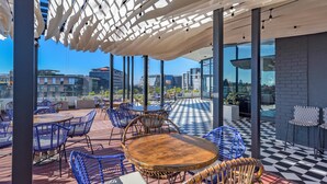 An outdoor seating area with round tables and blue chairs, a checkered floor, and a view of the cityscape through large windows.