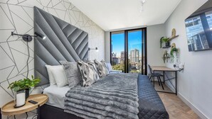 A modern bedroom with a geometric-patterned wall, a large bed with a grey quilt, a desk with a chair, and a view of the city through a large window.