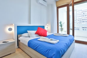 Ein modernes Hotelzimmer mit einem großen Bett, einem Nachttisch, einer Fernbedienung und Blick auf die Stadt durch ein großes Fenster.