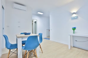 Ein moderner, gut beleuchteter Raum mit Esstisch, blauen Stühlen, einem kleinen Beistelltisch mit einer Vase und einer Klimaanlage an der Wand.
