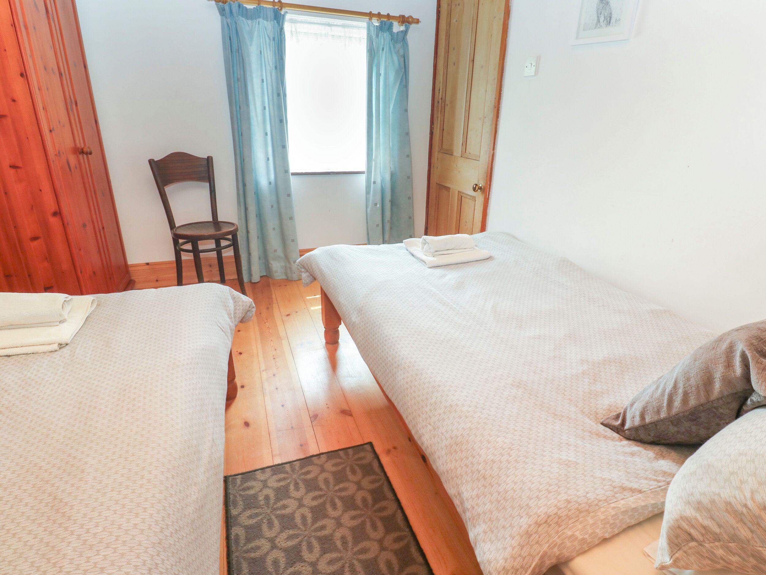 A room with two beds, wooden flooring, a chair, and a window with curtains.