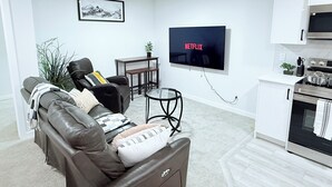 A modern living room with a leather sofa, a small coffee table, a TV mounted on the wall, and a kitchen area with a stove and cabinets.