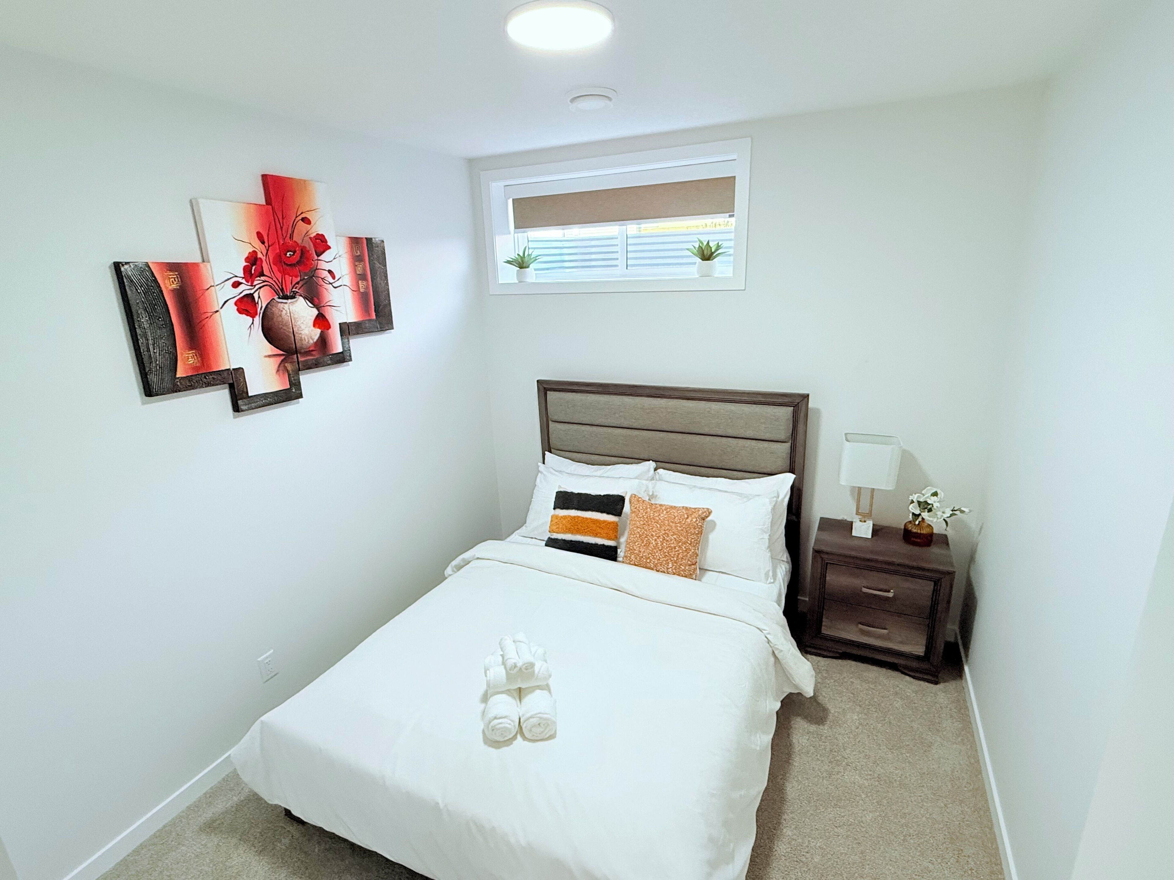 Une chambre à coucher bien rangée, avec un lit, une table de chevet, une fenêtre et des œuvres d’art accrochées au mur.