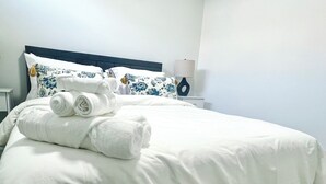 A neatly made bed with white linens and rolled towels, a dark headboard, and a bedside table with a lamp and decorative vase.