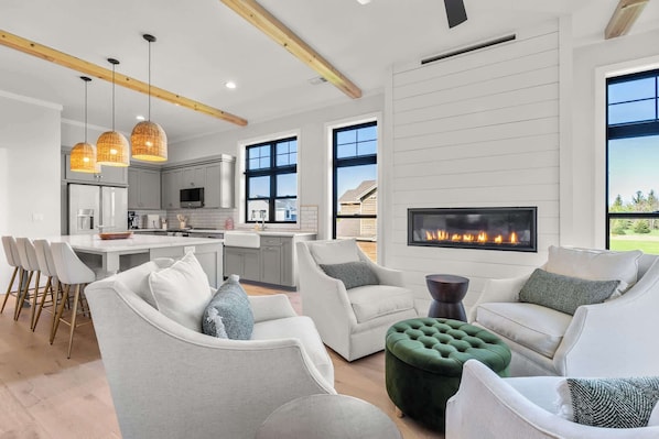 A modern living room with a fireplace, grey sofas, and a dining area with a kitchen in the background.