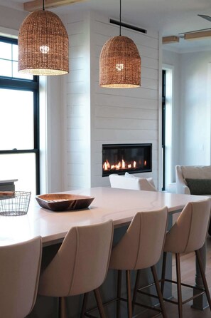 A modern dining area with a large table, six chairs, two hanging wicker pendant lights, and a fireplace.