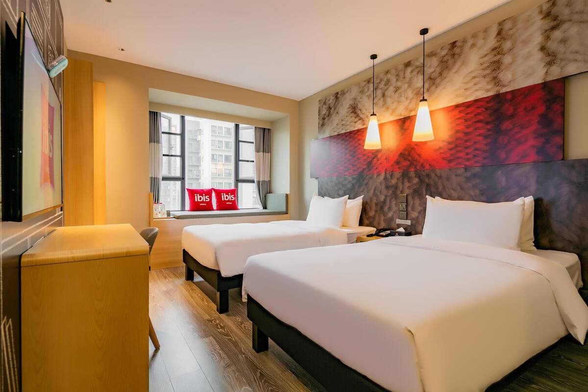 Habitación de hotel con dos camas, un escritorio de madera, una ventana con cortinas y una obra de arte colgada en la pared.