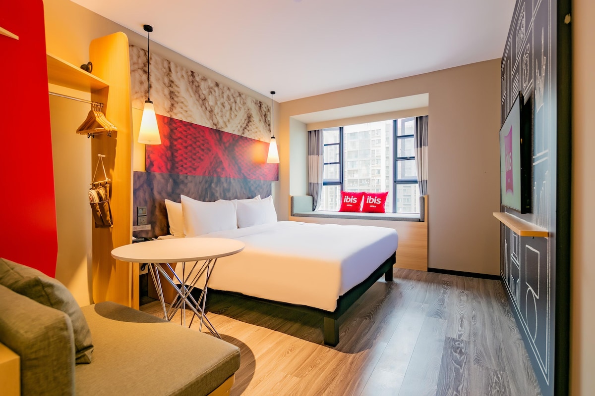 A hotel room with a bed, a small table, a chair, a window with curtains, and a wall with a patterned design.