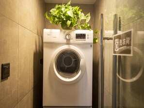 Un lave-linge à chargement frontal doté d’un panneau de commande, d’une plante en pot posée sur le dessus et d’une porte de douche en verre.