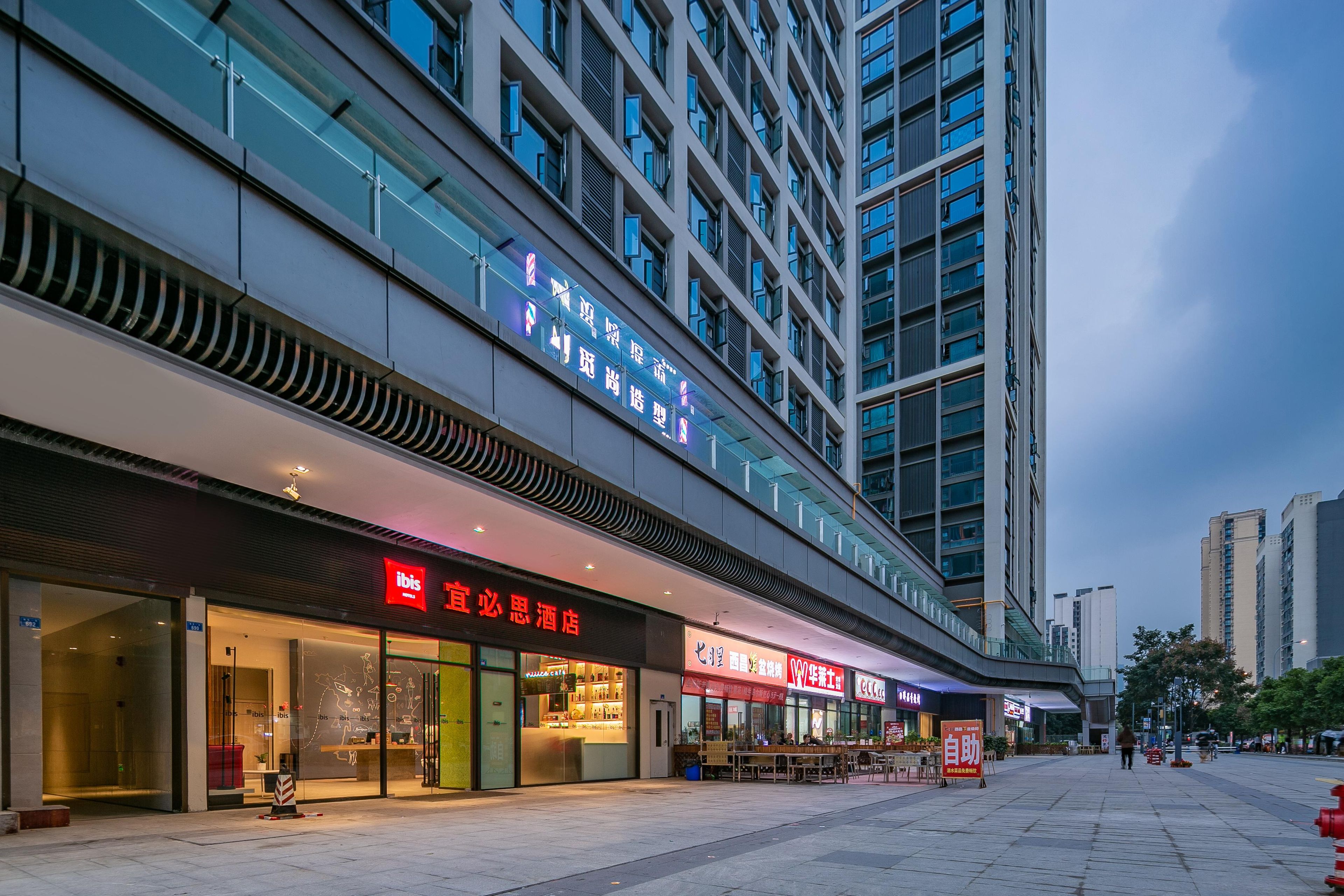 A modern hotel with a glass facade, outdoor seating area, and signage for amenities.