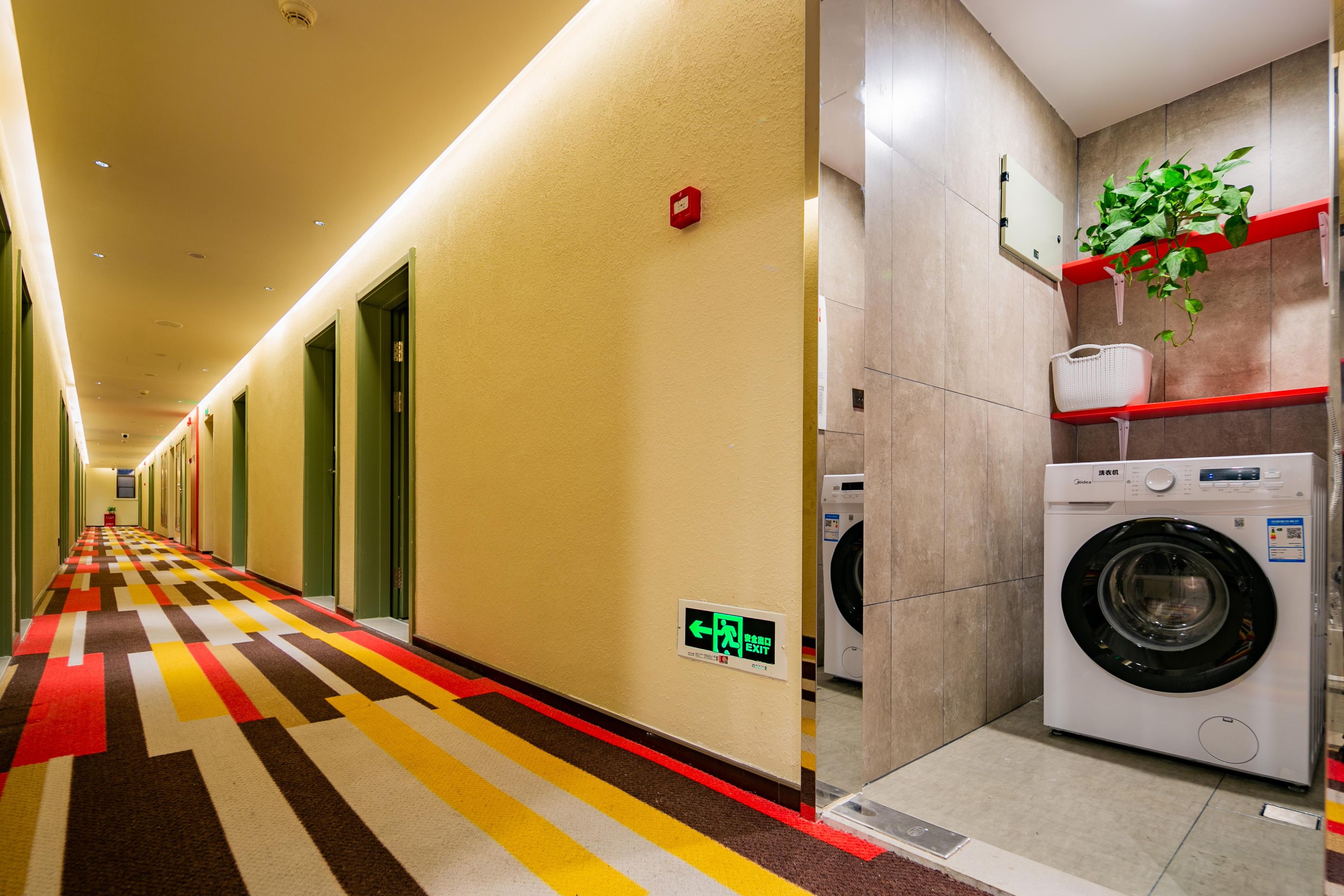 Corredor de hotel com carpete estampado, sinalização de saída vermelha e área de lavanderia com máquina de lavar e uma planta.