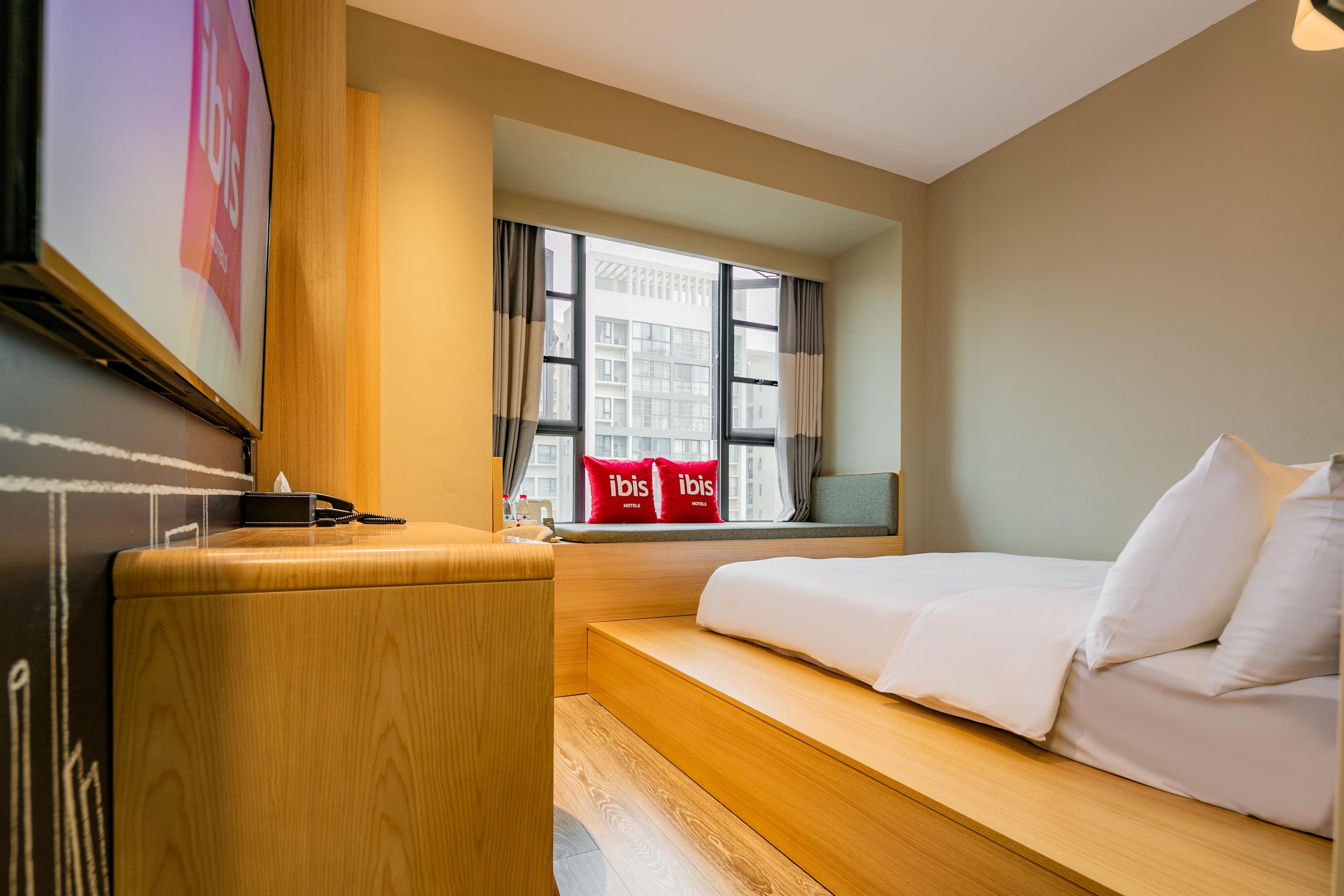 Quarto de hotel com cama, mesa de cabeceira de madeira, janela com cortinas e vista para edifícios.