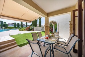 An outdoor dining area with a table set for two, surrounded by chairs, under a canopy.