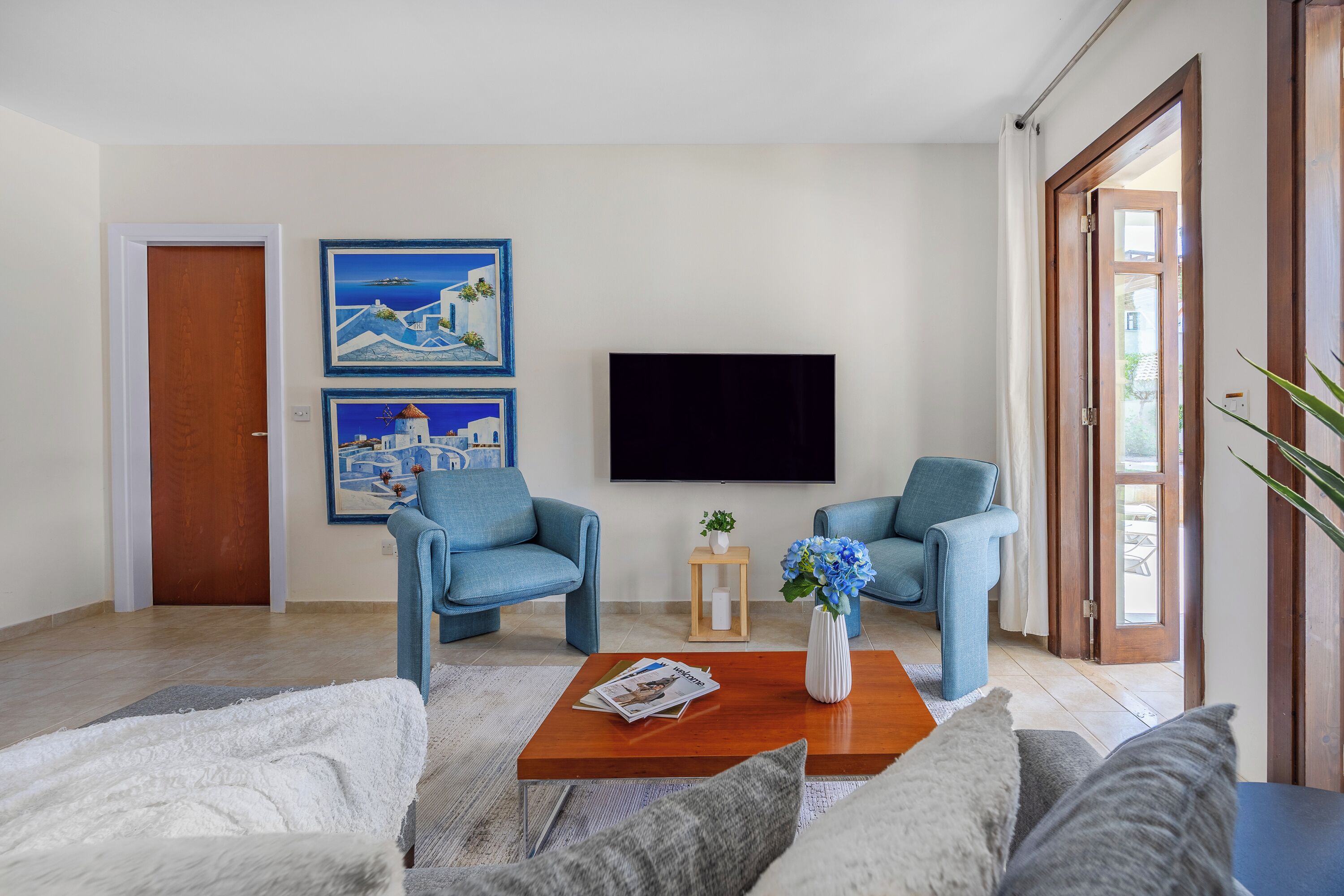 Een woonkamer met een houten salontafel, twee blauwe fauteuils, een witte bank en een flatscreen televisie die aan de muur is bevestigd.