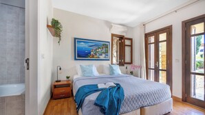 A bedroom with a large bed, wooden flooring, a nightstand, a bedside table, a wall-mounted plant, and a painting. - Da02 Adjacent to Aphrodite Hills Pool View (Kouklia)