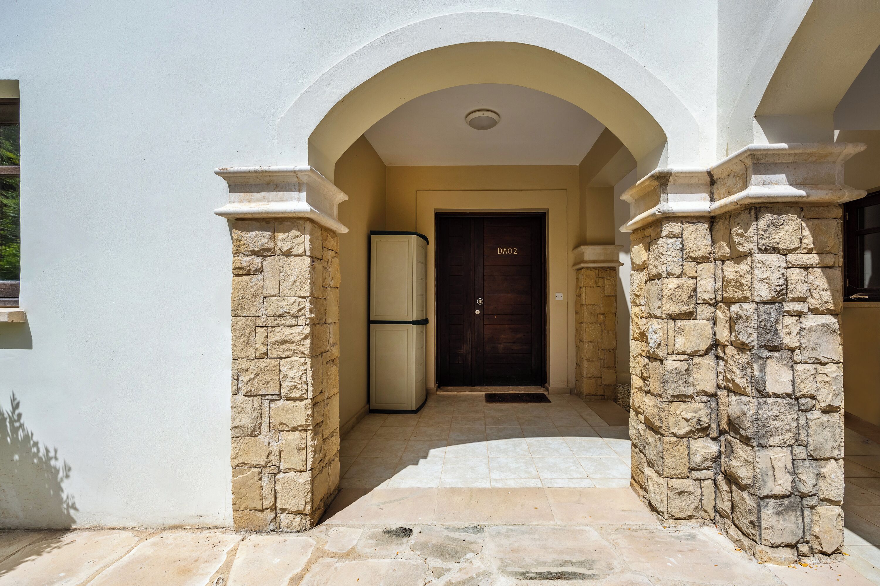 Une entrée en arc de pierre mène à un bâtiment. Une porte portant l’inscription « DAO2 » se trouve à droite, et un réfrigérateur est placé sur le côté gauche.