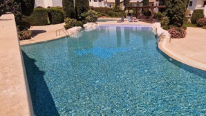 A clear swimming pool with a stone sculpture, surrounded by well-maintained landscaping and a covered area with lounge chairs. - Da02 Adjacent to Aphrodite Hills Pool View (Kouklia)