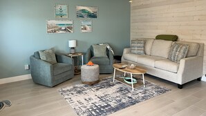 A living room with a grey sofa, a grey armchair, a round coffee table, a small ottoman, and a ceiling fan. There are wall-mounted picture frames and a light switch on the wall.