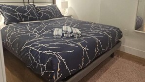 A bed with a dark blue and white leaf pattern bedspread, a headboard, a bedside lamp, and a framed picture on the wall.