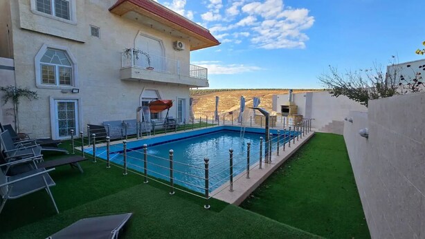 A swimming pool with a surrounding deck, a building with a balcony, and a clear sky. - Charming 3-bedroom scenic villa with a pool, WiFi in the lovely north (يبلا (إربد))