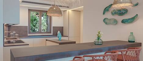 A modern kitchen with a central island, pendant lights, and wicker chairs.
