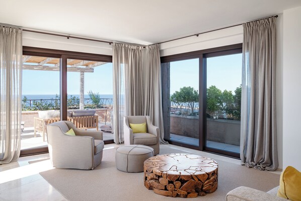 A modern living room with a sofa, armchair, ottoman, and coffee table, large windows with curtains, and a view of the outdoors. - La Tiara di Cervo (Arzachena)