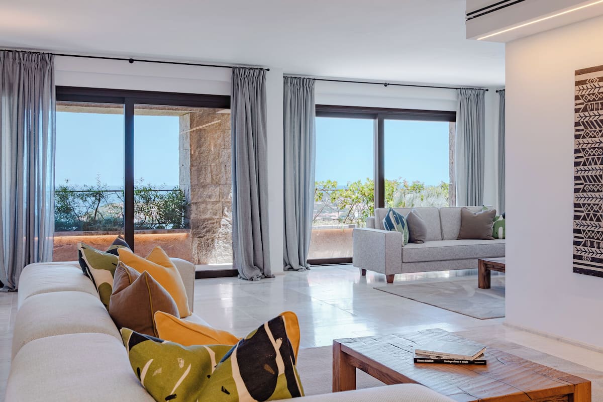 A modern living room with a sofa, a coffee table, and a couch. There are large windows with curtains, a stone wall, and a view of greenery.