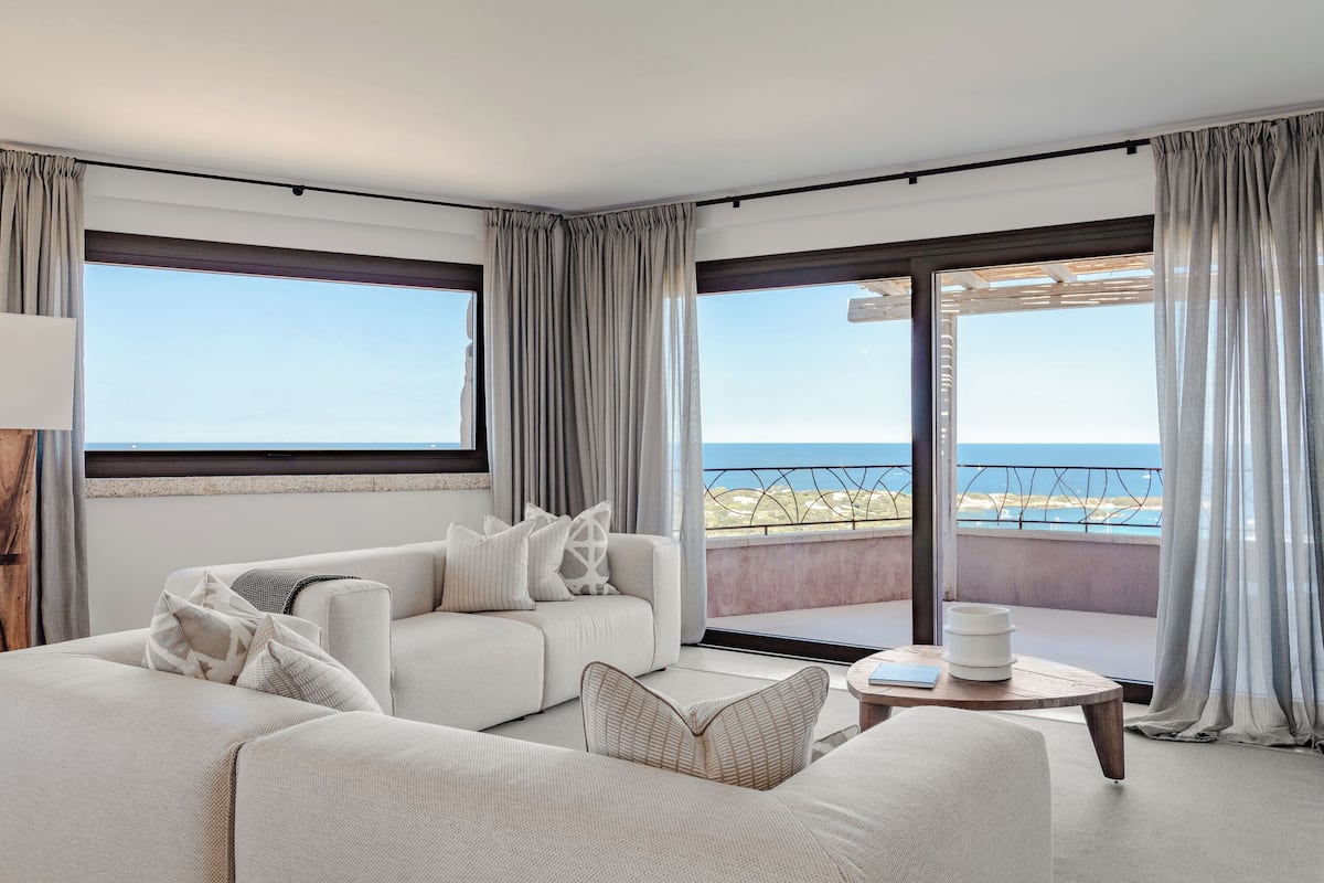 A modern living room with a large sofa, a coffee table, and a view of the sea through large windows.