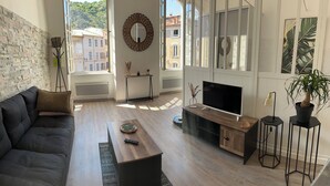 Ein modernes Wohnzimmer mit einer Couch, Fernseher, einem Holztisch und Blick auf Gebäude durch große Fenster.