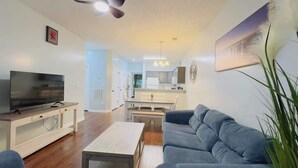 A modern living room with a flat-screen TV, a sofa, a coffee table, and a dining area in the background.