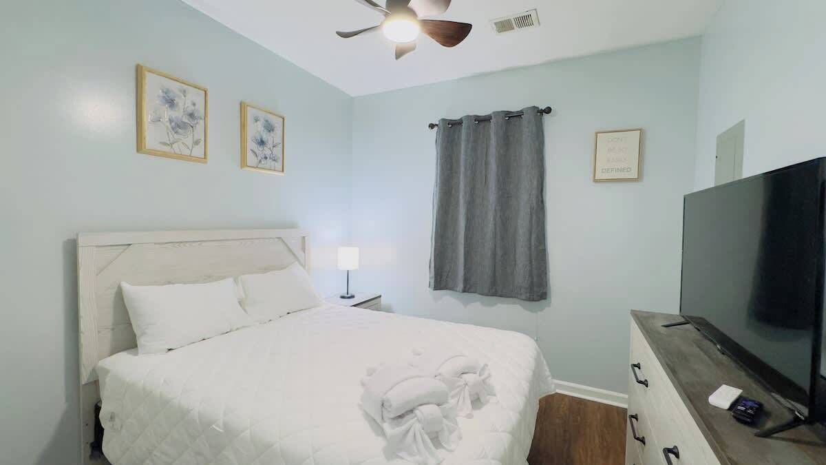 Une chambre à coucher avec un lit, un ventilateur de plafond, une télévision et une fenêtre avec des rideaux.