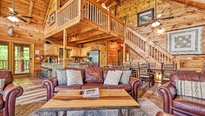 A spacious log cabin interior with a central living area featuring a wooden coffee table, leather sofas, and a staircase leading to an upper level.