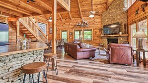A spacious log cabin interior with a stone fireplace, wooden floors, a bar area, and a cozy seating area.