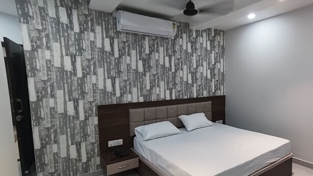 A bedroom with a modern design, featuring a patterned wall, a bed with white linens, a nightstand, and an air conditioning unit. - Hotel Shri Vishwanath (Varanasi)