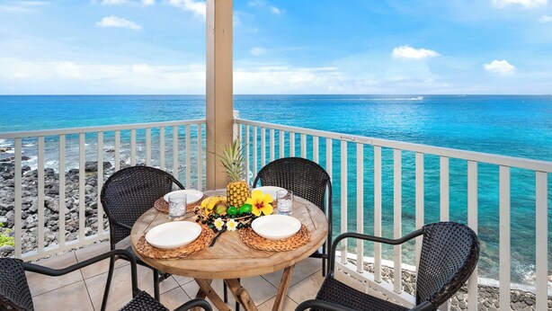 Une table en bois, des assiettes blanches, des chaises en osier noir et une corbeille de fruits tropicaux sur un balcon offrant une vue sur l’océan.