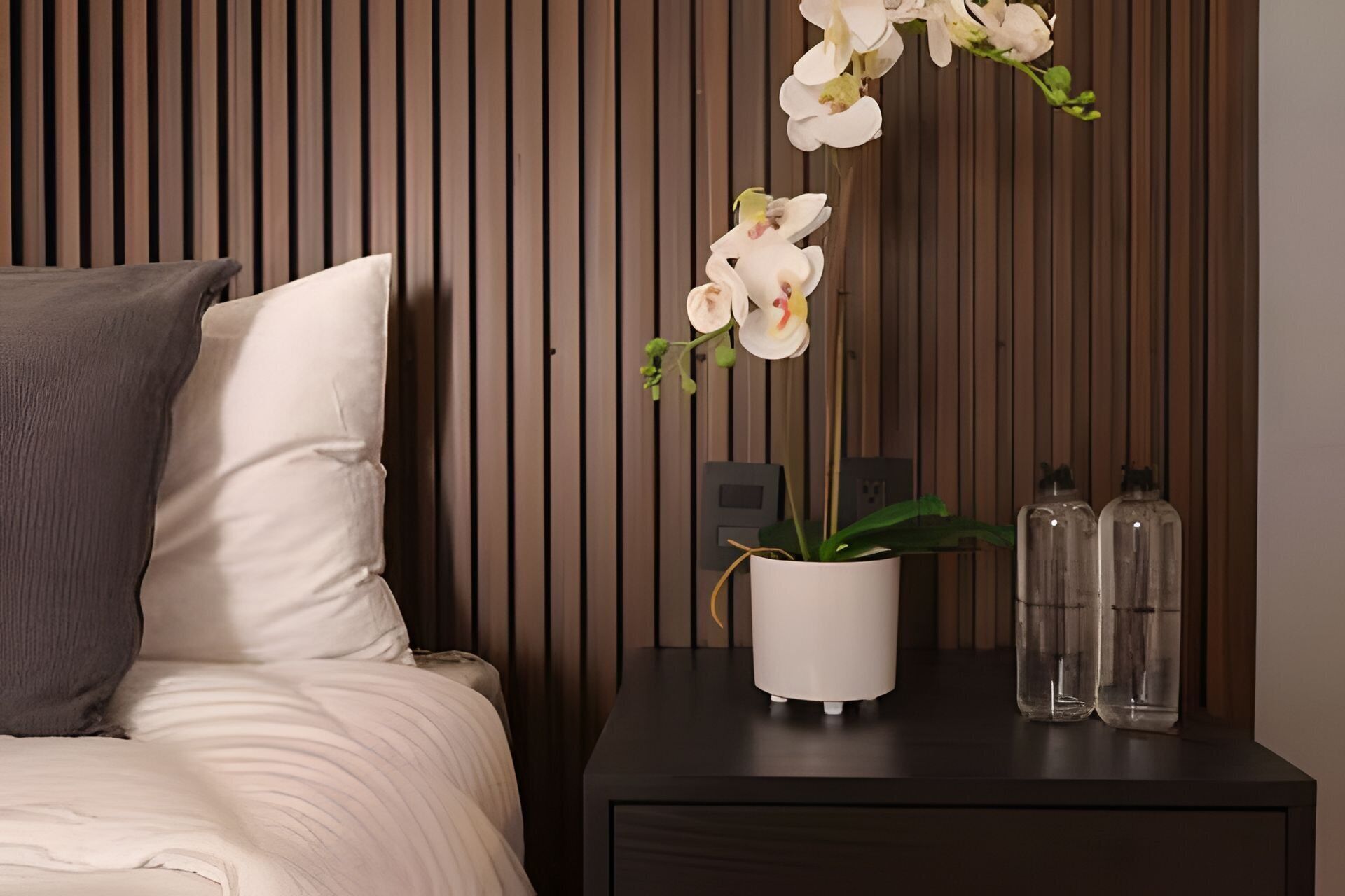 A bed with a white pillow and a dark nightstand with a white vase holding white orchids, two clear bottles, and a power outlet.