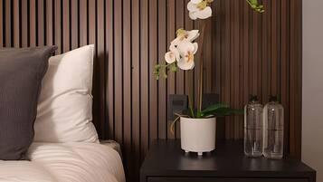 A bed with a white pillow and a dark nightstand with a white vase holding white orchids, two clear bottles, and a power outlet.